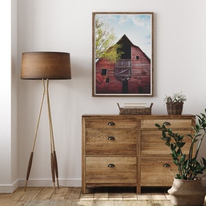 May include: A rustic wooden dresser with six drawers and black metal handles. A framed photograph of a red barn with a blue sky and green trees hangs above the dresser. A brown floor lamp with a brown lampshade stands to the left of the dresser. A potted plant with green leaves sits on the right side of the dresser.