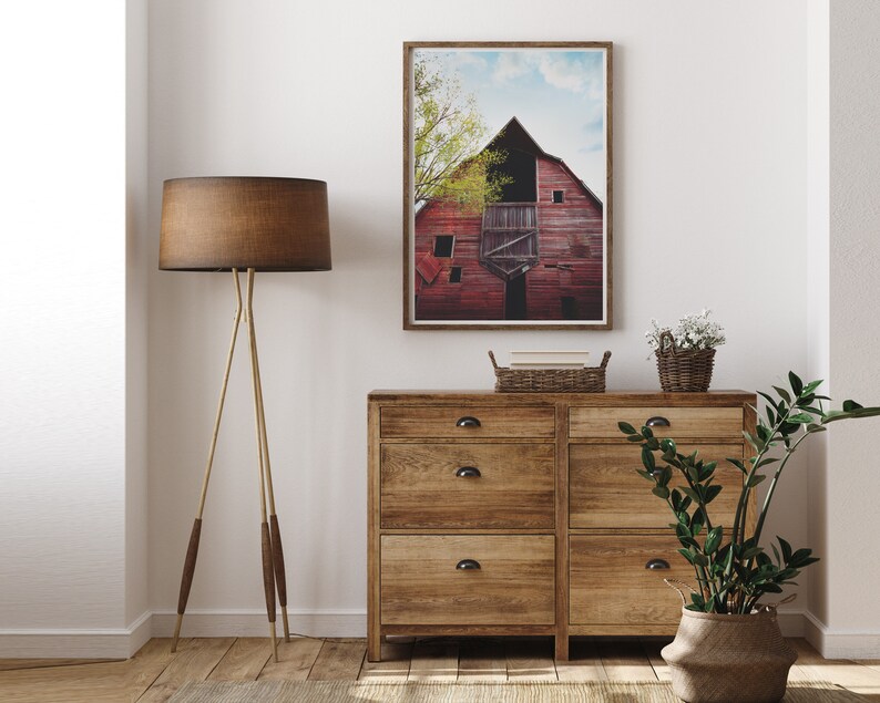 May include: A rustic wooden dresser with six drawers and black metal handles. A framed photograph of a red barn with a blue sky and green trees hangs above the dresser. A brown floor lamp with a brown lampshade stands to the left of the dresser. A potted plant with green leaves sits on the right side of the dresser.
