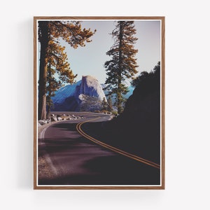 Pu&ograve; includere: Una strada tortuosa conduce a una vista di Half Dome, una formazione rocciosa di granito nel Parco Nazionale di Yosemite, in California. La strada &egrave; fiancheggiata da alberi e il cielo &egrave; di un azzurro chiaro.