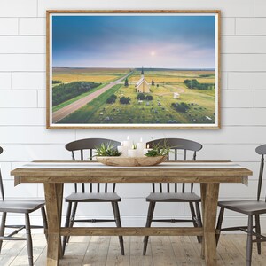 May include: An aerial photograph of a small, white church with a spire, surrounded by a graveyard. The church is situated in a rural area with a long, straight road leading to it. The sky is a light blue with some clouds.