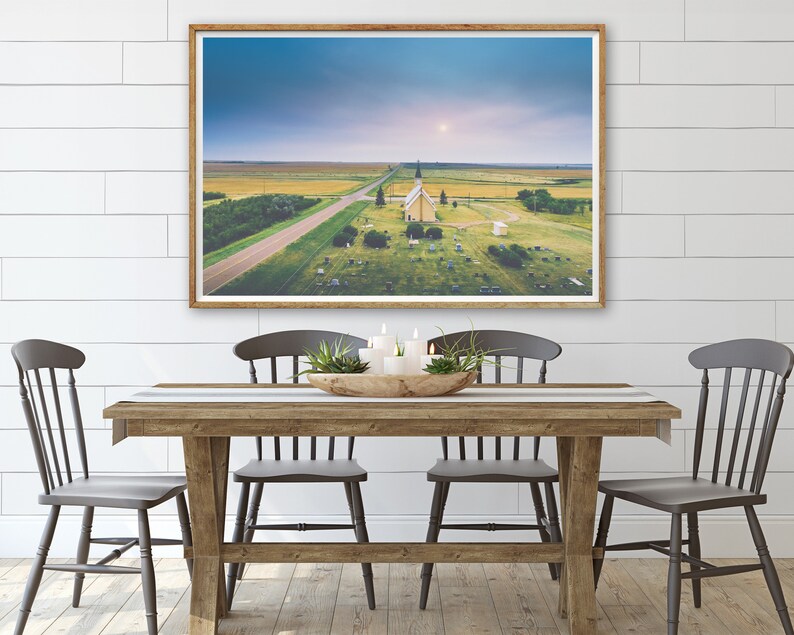 May include: An aerial photograph of a small, white church with a spire, surrounded by a graveyard. The church is situated in a rural area with a long, straight road leading to it. The sky is a light blue with some clouds.