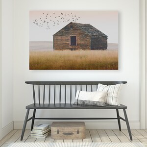 May include: A weathered wooden farmhouse with a faded brown roof sits in a field of tall, golden grass. A flock of birds flies overhead against a pale pink sky.