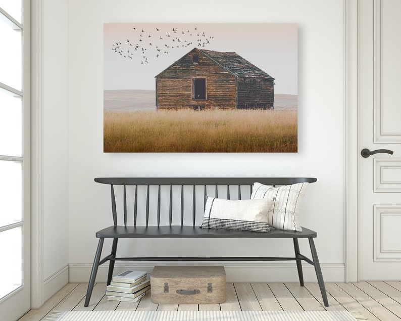 May include: A weathered wooden farmhouse with a faded brown roof sits in a field of tall, golden grass. A flock of birds flies overhead against a pale pink sky.