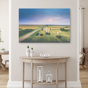 May include: An aerial view of a small, white church with a steeple in a grassy field. The church is surrounded by a cemetery and a dirt road leads to it. The sky is a light blue with a few clouds.