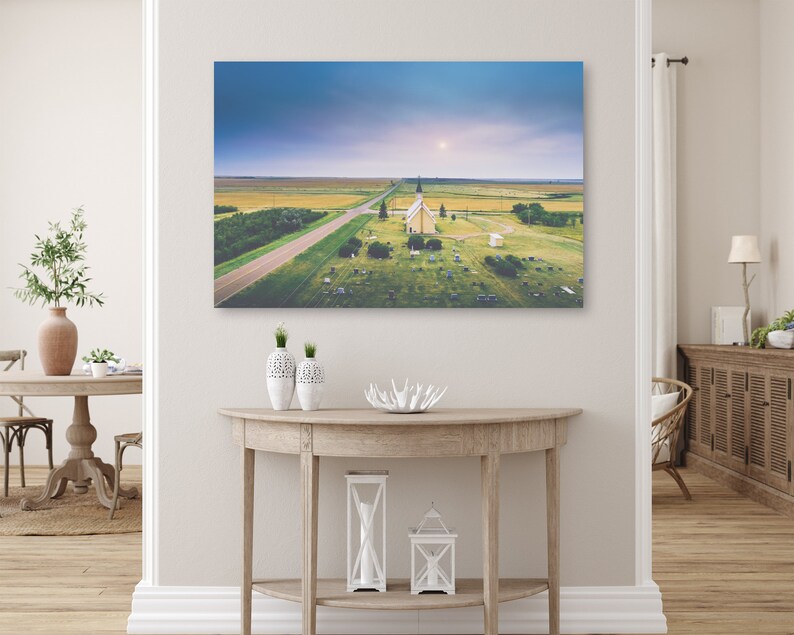 May include: An aerial view of a small, white church with a steeple in a grassy field. The church is surrounded by a cemetery and a dirt road leads to it. The sky is a light blue with a few clouds.