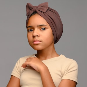 May include: A dark brown ribbed turban headwrap featuring a prominent bow detail on the front. The fabric has a textured, vertically striped appearance, and the headwrap is designed to fit snugly around the head with a gathered, pleated construction.