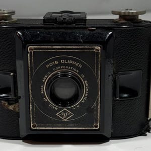 May include: A vintage black PD16 Clipper camera with a leather strap. The camera has a rectangular body with a lens in the center and dials on top. The camera's face has the text "PD16 CLIPPER CORPORATION IN BINGHAMTON N.Y." and the Agfa logo.