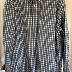 May include: A long-sleeved button-down shirt with a green, white, and navy blue plaid pattern. The shirt has a button-down collar, a chest pocket with a green logo, and paisley patterned cuffs. The shirt is made of a woven fabric.