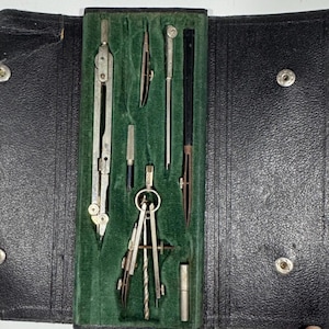 Puede incluir: Un juego de dibujo antiguo en un estuche de cuero negro. El juego incluye varios compases, divisores y otras herramientas de dibujo de color plateado, dispuestos sobre un forro de terciopelo verde. El estuche se cierra con cierres a presión.