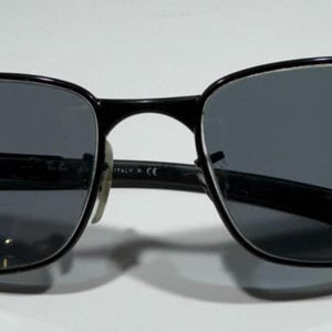 Puede incluir: Gafas de sol negras con lentes de color gris oscuro. Las monturas son rectangulares con bordes redondeados y un puente fino. Las patillas también son negras y tienen una ligera curva. Las gafas están sobre un fondo blanco.