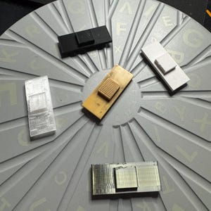May include: Five different 3D printed keycaps in various colors, including black, white, beige, and gray. The keycaps are arranged on a circular surface with a pattern of letters and numbers.