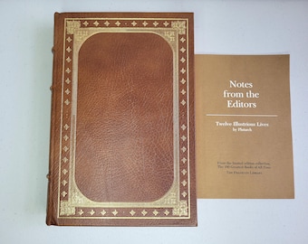 Può includere: Un libro rilegato in pelle marrone con dettagli in foglia d'oro sulla copertina, accompagnato da un opuscolo color marrone chiaro intitolato Notes from the Editors, Twelve Illustrious Lives by Plutarch, dalla collezione limitata The 100 Greatest Books of All Time.