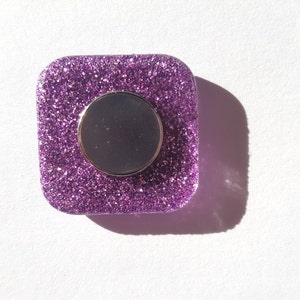 Pu&ograve; includere: Un magnete quadrato di glitter viola con un centro rotondo argentato.
