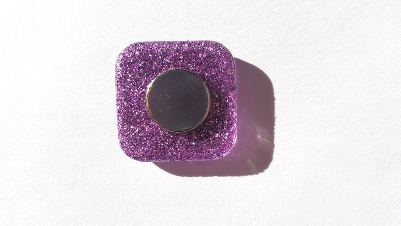 Pu&ograve; includere: Un magnete quadrato di glitter viola con un centro rotondo argentato.