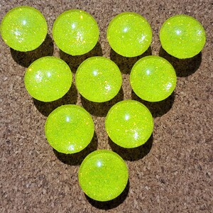May include: A group of ten round, yellow, glitter-covered, decorative ornaments arranged in a heart shape on a brown corkboard.