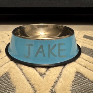May include: A blue pet food bowl with a silver rim and the name "JAKE" printed on the front.