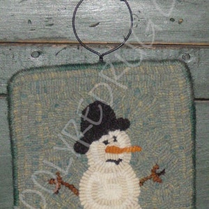 May include: A hand-hooked, square wall hanging featuring a snowman design. The snowman has a black top hat, an orange carrot nose, and twig arms. The background is a muted teal color, and the snowman is made of off-white yarn. A black metal hanger is attached.