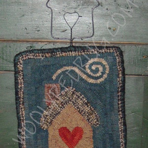 May include: A handcrafted, rectangular wall hanging with a teal background and a house design. The house features a tan body, a red heart, and a patterned roof. A wire house-shaped hanger with a green knob is attached.