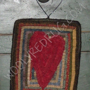 May include: A handcrafted, square rug with a large red heart in the center. The rug features a multi-colored, striped border in shades of brown, blue, green, and yellow. A black metal heart-shaped hanger is attached to the top.