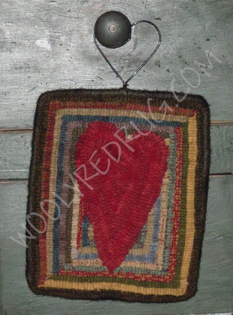 May include: A handcrafted, square rug with a large red heart in the center. The rug features a multi-colored, striped border in shades of brown, blue, green, and yellow. A black metal heart-shaped hanger is attached to the top.