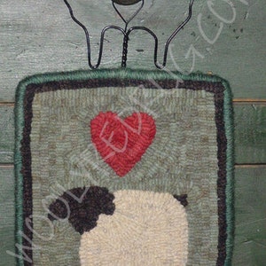 May include: A handcrafted, square wall hanging featuring a sheep design. The wool art piece has a green border and a red heart above the sheep. A black metal sheep silhouette and a green knob are attached at the top.