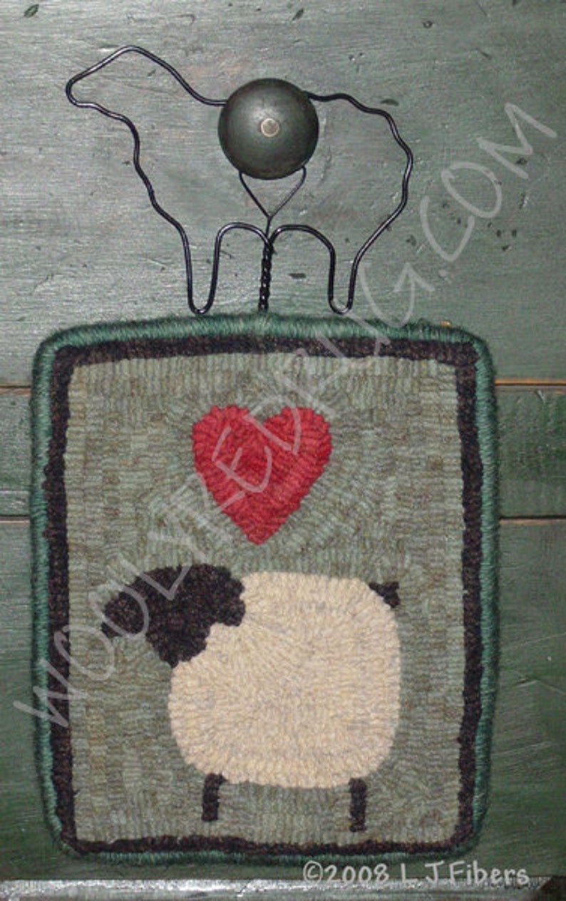 May include: A handcrafted, square wall hanging featuring a sheep design. The wool art piece has a green border and a red heart above the sheep. A black metal sheep silhouette and a green knob are attached at the top.