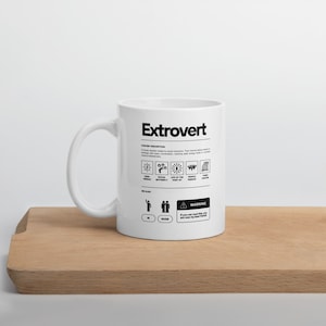 Op de afbeelding: Witte keramische mok met een zwart-wit grafisch ontwerp met de tekst "Extrovert" en een definitie van een extravert. De mok heeft een zwart handvat.