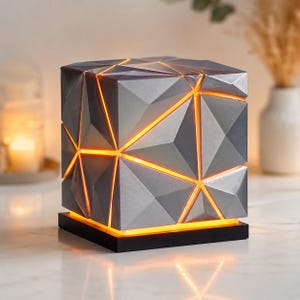 Peut inclure: Lampe grise en forme de cube géométrique, avec une base noire. La surface de la lampe est composée de facettes triangulaires, avec une lumière orange chaude émanant des espaces. Un élément de décoration intérieure moderne.