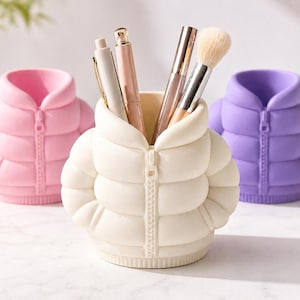 Könnte beinhalten: Drei Stifthalter in Form von Pufferjacken in Rosa, Creme und Lila. Der cremefarbene Halter enthält Stifte und Make-up-Pinsel. Jeder Halter hat einen Reißverschluss und einen runden Kragen. Die Halter sind aus einem glatten, festen Material.