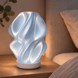 Puede incluir: Una lámpara de noche moderna, de color azul claro, con un diseño único y ondulado. La lámpara se apoya sobre una base de madera y emite un brillo suave. La lámpara está colocada sobre una mesita de noche de madera junto a una cama con almohada y manta blancas.