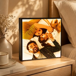 May include: A black square photo frame displaying a backlit image of three people smiling. The frame sits on a wooden nightstand next to a vase of white flowers and a cup and saucer.