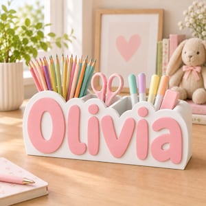 Könnte beinhalten: Ein weißer Schreibtisch-Organizer mit dem Namen Olivia in erhabenen rosa Buchstaben. Der Organizer verfügt über mehrere Fächer für Buntstifte, eine rosa Schere, Marker und einen rosa Radiergummi. Das Design ist abgerundet und wolkenartig.
