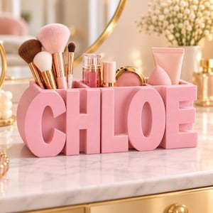 Op de afbeelding: Een lichtroze bureau-organizer in de vorm van de naam CHLOE, met meerdere vakken voor diverse make-upkwasten, lipgloss-tubes, een compacte blush en een roze cosmeticaspons. De organizer heeft een gladde, matte afwerking.