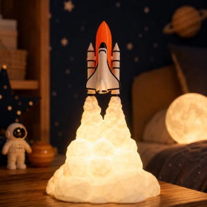 Peut inclure: Lampe en forme de fusée blanche, orange et noire, avec une base en forme de nuage lumineux. La lampe est conçue pour ressembler à une fusée au décollage. L'arrière-plan comprend un ciel étoilé et une lampe en forme de lune.