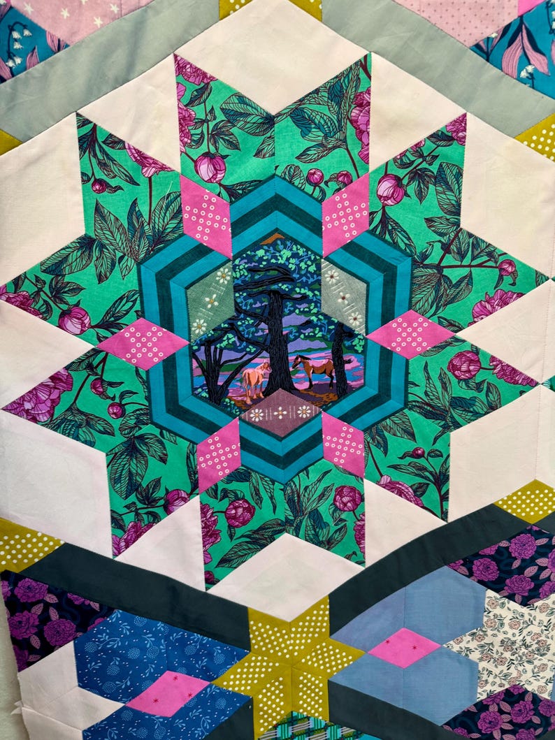 May include: Close-up of a handmade quilt with a starburst design. The quilt features a central star with a landscape scene, surrounded by teal, pink, and green floral patterns. White and geometric shapes complete the design.