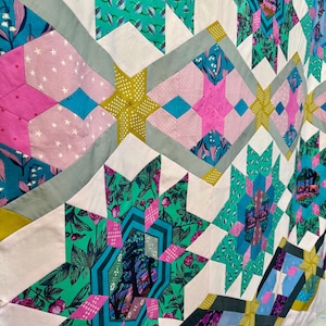 May include: A colorful quilt with a star pattern. The quilt features various fabrics in shades of pink, teal, green, and yellow. The star shapes are outlined with gray fabric. The quilt has a patchwork design.
