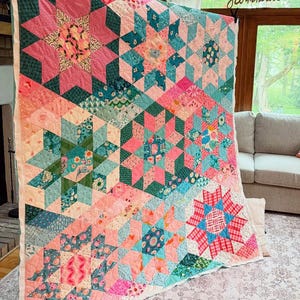 Puede incluir: Un edredón grande, hecho a mano, con un diseño de estrella en tonos rosa, verde azulado y verde. El edredón presenta formas geométricas y patrones florales, creando una estética vibrante y acogedora. El edredón se exhibe en una pared.