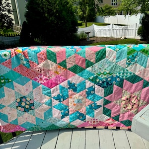Op de afbeelding: Een kleurrijke, handgemaakte quilt met een sterrenpatroon. De quilt heeft roze, turquoise en groene stoffen. De quilt wordt buiten op een wit houten oppervlak gepresenteerd.