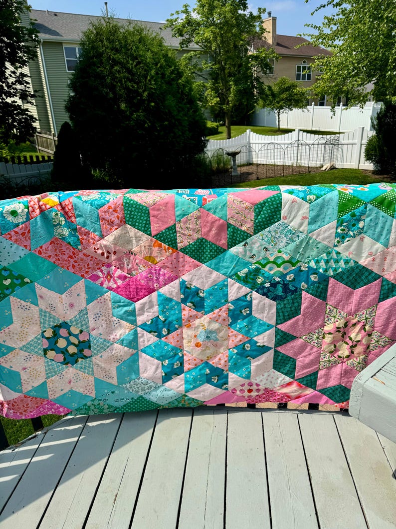 Op de afbeelding: Een kleurrijke, handgemaakte quilt met een sterrenpatroon. De quilt heeft roze, turquoise en groene stoffen. De quilt wordt buiten op een wit houten oppervlak gepresenteerd.