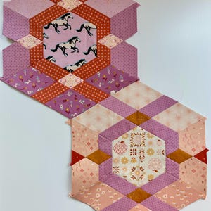 May include: Two handmade quilted hexagon shapes. One features a pink background with running horses, and the other has a floral pattern. The quilts are in shades of pink, orange, and purple.