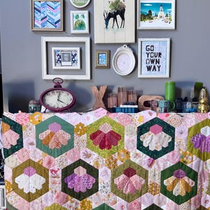 May include: A handmade quilt featuring a repeating pattern of large hexagons, each containing a floral-inspired design made of smaller fabric segments in shades of pink, green, yellow, and purple, set against a background of light pink floral fabric.