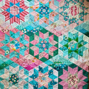 Op de afbeelding: Een kleurrijke patchwork quilt met een geometrisch sterrenpatroon. De quilt heeft verschillende stoffen in roze, teal en groene tinten. De stervormen zijn samengesteld uit kleinere ruit- en driehoekstukken, waardoor een textuurrijk ontwerp ontstaat.