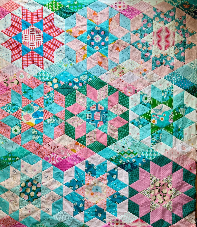 Op de afbeelding: Een kleurrijke patchwork quilt met een geometrisch sterrenpatroon. De quilt heeft verschillende stoffen in roze, teal en groene tinten. De stervormen zijn samengesteld uit kleinere ruit- en driehoekstukken, waardoor een textuurrijk ontwerp ontstaat.