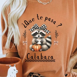 Może przedstawiać: Pomarańczowa koszulka z grafiką szopa pracza w kokardce i kubkiem, z dynią. Tekst brzmi "¿Que te pasa? Calabaza". Koszulka jest w stylu casual.