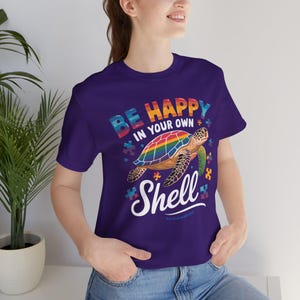 May include: A purple t-shirt featuring a colorful turtle with a rainbow shell. The shirt has the text "BE HAPPY IN YOUR OWN Shell" in a playful font. The design includes puzzle piece accents.