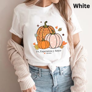 May include: White t-shirt featuring a fall-themed graphic with three pumpkins in orange, pink, and leopard print. The design includes leaves, hearts, and the text "Fe, Esperanza y Amor" in Spanish.