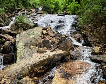 Könnte beinhalten: Ein Wasserfall stürzt über mit Moos bedeckte Felsen in einem üppigen grünen Wald. Das Wasser stürzt über das felsige Gelände und erzeugt eine ruhige und malerische Szene.