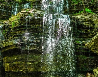 Könnte beinhalten: Ein Wasserfall stürzt über mit Moos bewachsenen Felsen in einem üppigen, grünen Wald. Das Wasser ist klar und glitzernd, und das umliegende Grün ist lebendig.