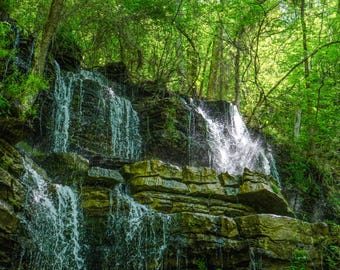 Könnte beinhalten: Ein malerischer Wasserfall stürzt über geschichtete Felsformationen. Das Wasser fließt über die Felsen und erzeugt ein erfrischendes Naturschauspiel. Üppige grüne Bäume umgeben den Wasserfall und tragen zur natürlichen Schönheit bei.