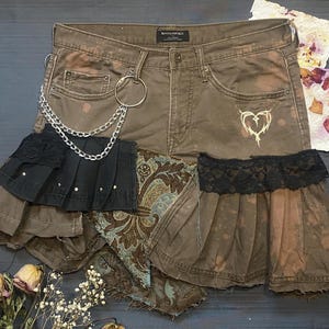 May include: Brown denim shorts with a layered skirt design. The shorts have a silver chain and ring detail, a heart graphic, and a black lace trim. Additional fabric layers include paisley and pleated black fabric.